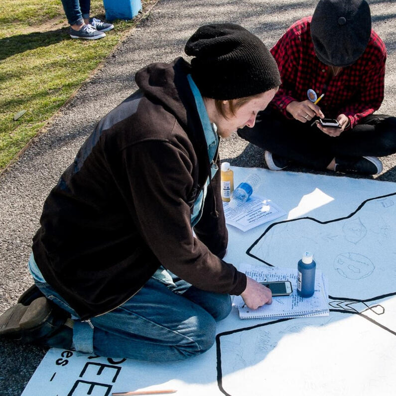 Jonathan kneeling on the floor over white paper that is being drawn on (someone else caught in the picture kneeling without a head). Jonathan is wearing a black droopy hat over straight blonde hair and wearing a black sweater, blue scarf, and blue jeans